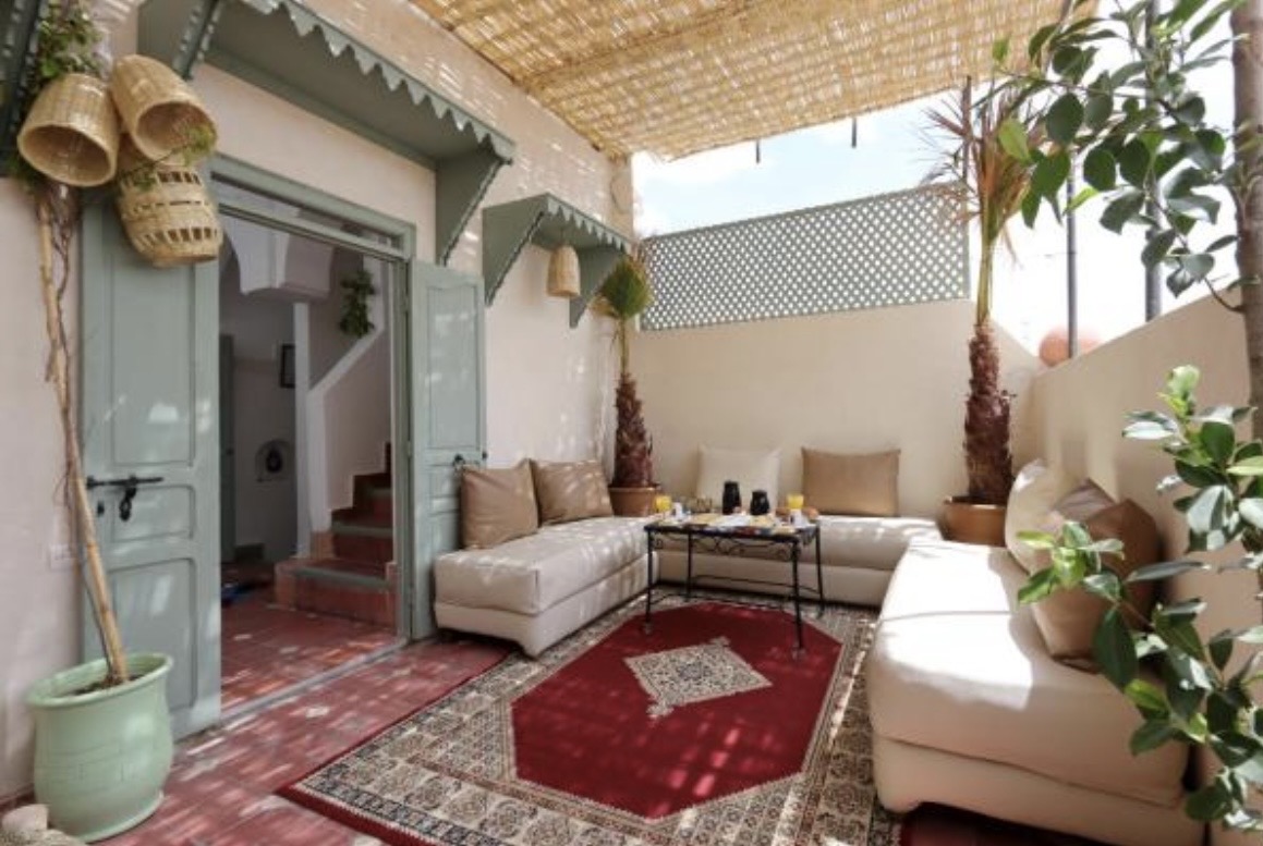 Terrasse d'un riad à Marrakech : banquettes crème, portes vert sauge et tapis rouge